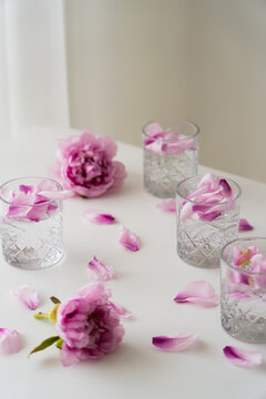 crystal glasses with tonic and floral petals near pink peonies on white tabletop and grey background.