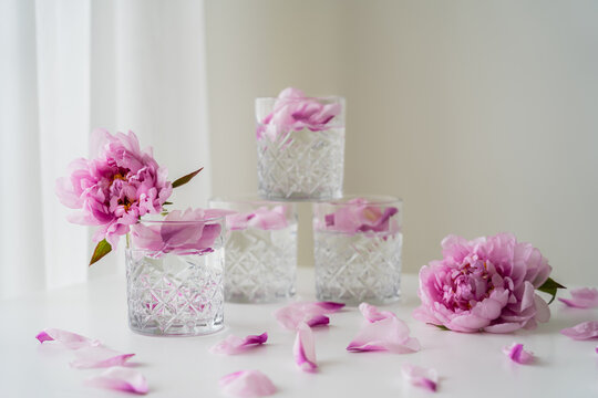 faceted glasses with tonic near pink peonies on white surface and grey background.