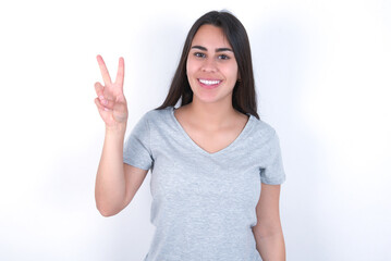 Young beautiful brunette woman wearing grey T-shirt over white wall showing and pointing up with fingers number two while smiling confident and happy.