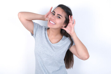 young beautiful brunette woman wearing grey t-shirt over white wall Trying to hear both hands on ear gesture, curious for gossip. Hearing problem, deaf