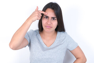 young beautiful brunette woman wearing grey t-shirt over white wall pointing unhappy at pimple on forehead, blackhead  infection. Skincare concept.