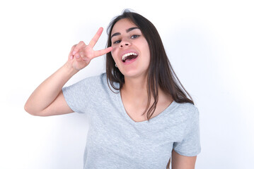 young beautiful brunette woman wearing grey t-shirt over white wall Doing peace symbol with fingers over face, smiling cheerful showing victory