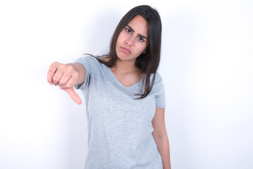 young beautiful brunette woman wearing grey t-shirt over white wall feeling angry, annoyed, disappointed or displeased, showing thumbs down with a serious look