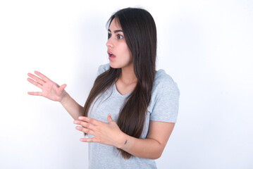 young beautiful brunette woman wearing grey t-shirt over white wall shouts loud, keeps eyes opened and hands tense.