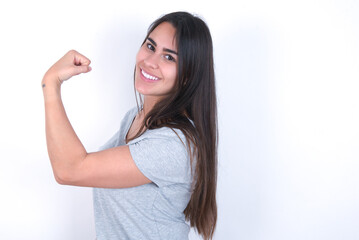 Portrait of powerful cheerful Young beautiful brunette woman wearing gray T-shirt over white wall showing muscles.