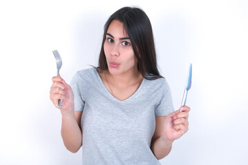 hungry Young beautiful brunette woman wearing gray T-shirt over white wall holding in hand fork knife want tasty yummy pizza pie