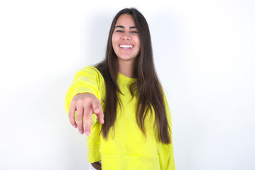 Excited positive Young beautiful brunette woman wearing yellow hoodie over white wall points index finger directly at you, sees something very funny. Wow, amazing