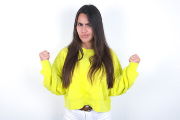 Irritated Young beautiful brunette woman wearing yellow hoodie over white wall blows cheeks with anger and raises clenched fists expresses rage and aggressive emotions. Furious model