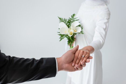 cropped view of bride with bouquet holding hands with blurred groom isolated on grey.