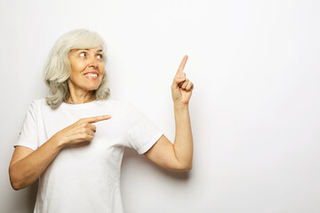 Elderly woman in t-shirt demonstrate, looking, pointing with fingers up to copy space standing over gray background