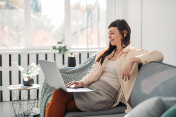 A young woman works on a laptop, communicates online in a modern bright office on a sofa on a sunny day.