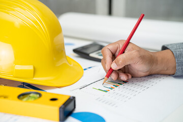 Asian engineer with plan project blueprint, graph and yellow helmet and engineering construction tools.