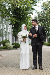 full length of happy muslim bride in wedding hijab and white dress holding bouquet near groom...