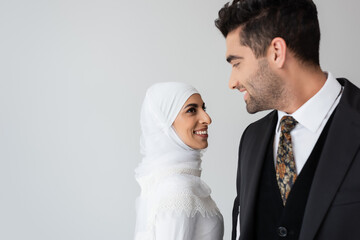 happy woman in wedding dress and hijab standing near joyful husband in suit isolated on grey.