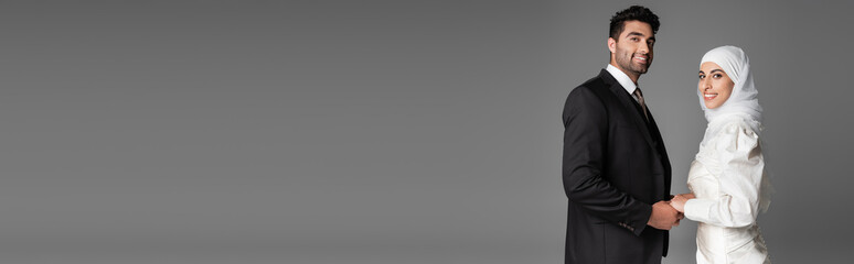 happy muslim bride in wedding dress and groom in suit holding hands isolated on grey, banner.