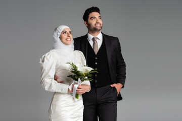 groom in suit posing near muslim happy bride with wedding bouquet on grey.