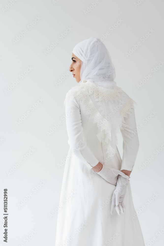 Wall mural muslim bride in gloves and white dress standing with hands behind back isolated on grey.