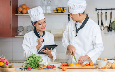 Two Asian professional couple chef wearing white uniform, hat, preparing ingredients, cooking meal in kitchen together, using tablet for recipe, smiling with happiness and confidence. Food Concept.