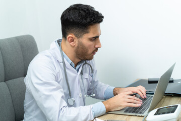 Young Caucasian male doctor in white medical uniform with a stethoscope around his neck sitting at a working desk, looking at the screen while typing and working with laptop in clinic examination room