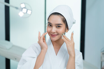 Asian woman in bathrobe and towel on her head gently touching skin on her face and smiling to be satisfied with herself a beauty while looking at camera like a mirror in beauty clinic's operating room