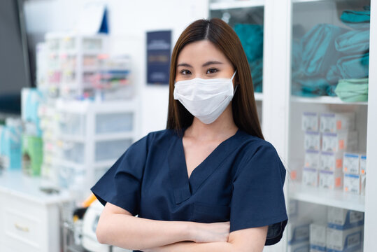 Young Beautiful Long-haired Asian Female Nurse Or Doctor In A Blue Uniform Wearing A Face Mask For Protective Covid-19 Standing With Arms Folded, Looking And Smiling At Camera Inside A Hospital Room.