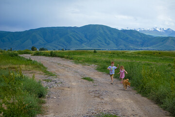 children and a dog run along a country road against the background of blue mountains