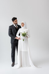 muslim groom hugging cheerful bride in wedding dress with bouquet on grey.