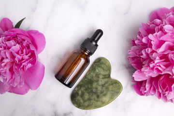 Glass bottle with oil, jade Gua sha stone for face massage on marble background with peony flowers