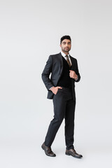 full length of muslim groom in formal wear standing with hand in pocket on grey.