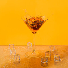 Martini cocktail glass with splashing alcohol drink and melting ice cubes against orange yellow...