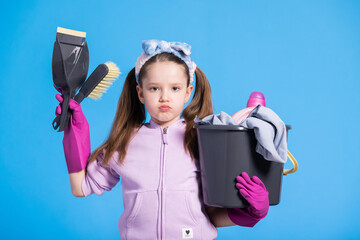 Disgruntled grumpy angry little girl in two ponytails with headband holds cleaning accessories in...