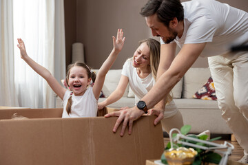 Excited young family having fun on moving day, happy parents pushing cardboard boxes with funny sweet little girl daughter riding in containers, looking at the camera and laughing about moving in.