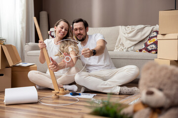 A happy couple in love are sitting on the floor of their new apartment, assembling living room furniture, the man holding a lampstand spinning it like a car steering wheel, fooling around.