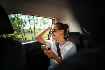 Young woman with a smartphone in her hands while sitting in a car in the back seat fastened with a...