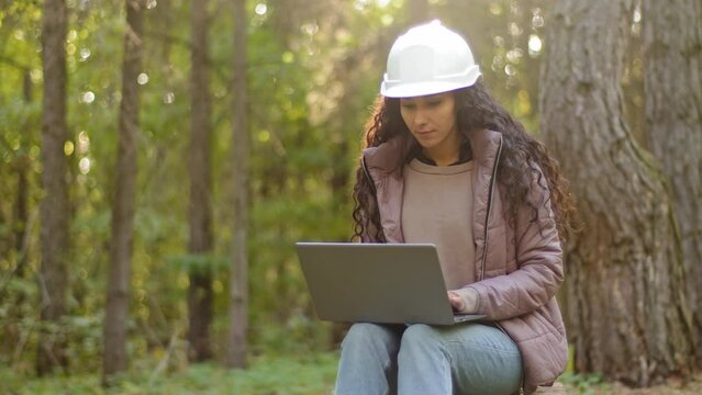 Attractive woman forestry engineer in protective helmet enters data into laptop takes reforestation action young experienced female specialist ecologist technician watching nature reserve checks trees