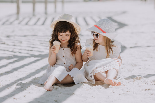 Beautiful little girls in white dress and hat eating ice cream on beach in summer time