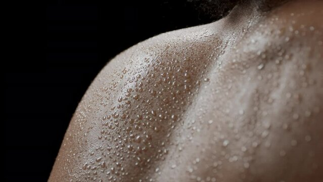 Big close-up shot of young good-looking slim African American female model with dark curly hair moves her wet shoulders on black background | Clean body concept