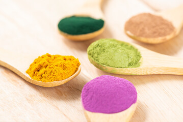 Multicolored superfood powders in spoons: sweet potato, matcha, spirulina, hemp, turmeric, carob on a wooden board.