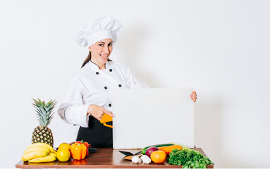 Chef woman with vegetables at table holding blank menu. Girl chef in the kitchen showing a blank board, Beautiful woman chef with table of vegetables holding a blank board