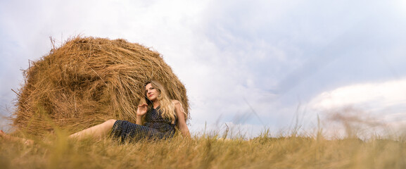 Girl and haystack. Thoughtful blonde woman in a polka-dot dress sits on the grass next to a haystack on the Sunset. Harvest concept. Banner