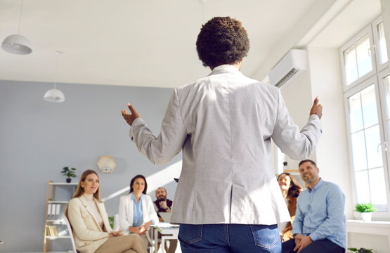 Female Business Coach And Talented Speaker Standing In Modern Office And Talking To Group Of People. Backside View From Behind Of Black Woman Meeting Team Of Employees And Making Motivational Speech