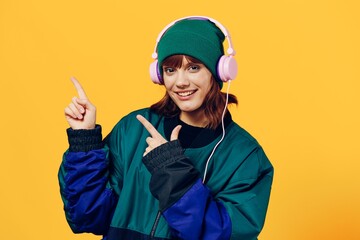 a cheerful woman in a green jacket and a hat in the style of the 90s enjoys music standing in pink headphones on a yellow background pointing her fingers at the empty space of the background