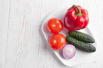 Vegetables lie on a plate on a white wooden background. Pepper, tomato, onion, cucumber, olives.
