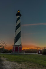 Fototapeta premium Vertical photograph of Hatteras Lighthouse at sunset mostly yellow and orange light in background blue hour sky. Light on in lighthouse green grass a white fence on Outer Banks Buxton North Carolina
