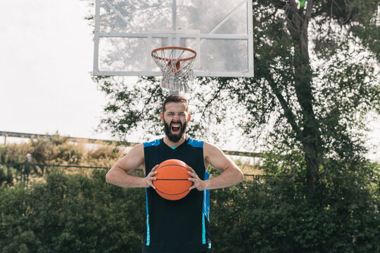 Young boy with a beard and shouting with joy under the basket after a street basketball game. Frontal image of a sporty man with a sleeveless shirt and an orange ball in his hands. Sport conceptring