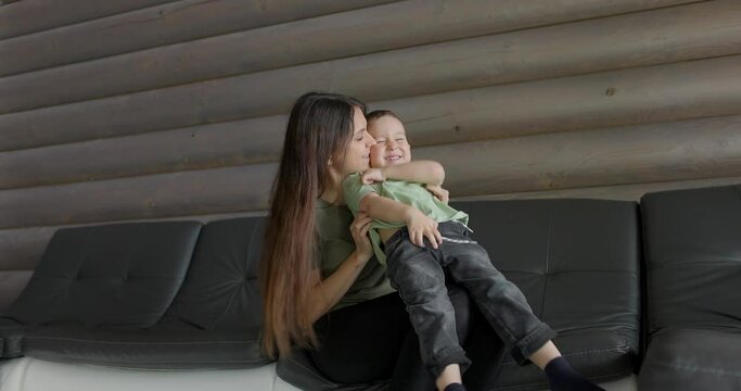 Little cheerful son sit on mum laps enjoy funny active playtime, laughing mom play tickle small boy sit on sofa in living room