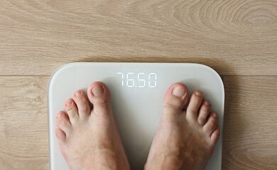 Man weighing himself - male bare feet stepping on white digital floor scales at home: close up view. Measuring weight, control, wellness and diet concept