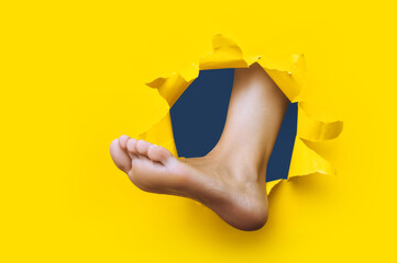 A left woman's foot appears in a torn hole in yellow paper. Healthy legs concept. Joke. Background...