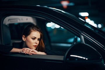  photo from the side, at night, of a woman sitting in car and looking out of the window looking into the side view mirror with her hands on the edge of the car with a thoughtful expression on her face