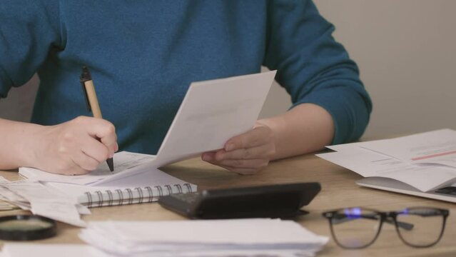 female woman hands in blue shirt looking through paper documents, utility bills at home, signing, sorting checks or vouchers. home budget, counting concept. glasses, calculator on desk table. working